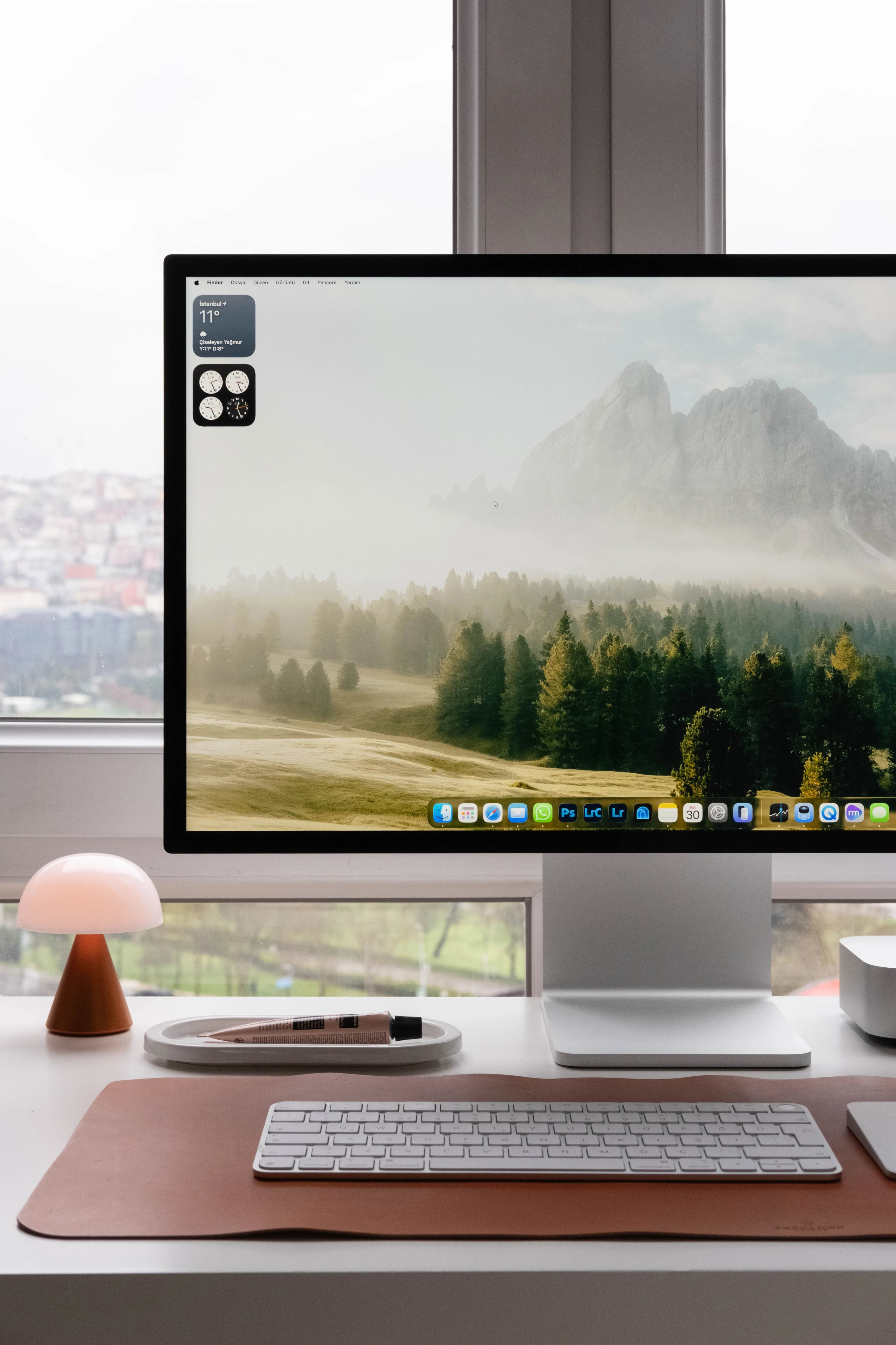 A tidy studio desk by a window: a large display, keyboard, and small lamp