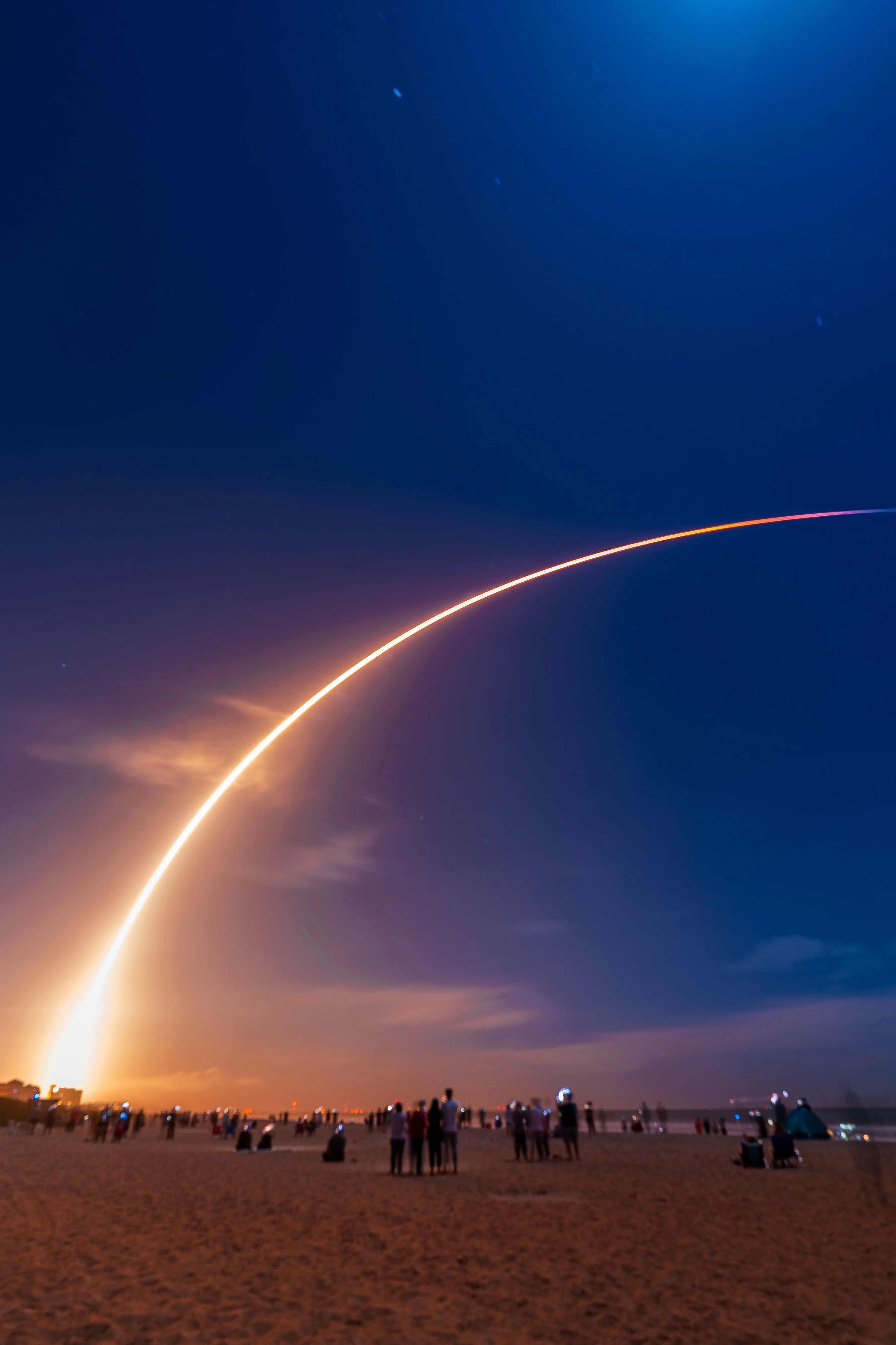 A rocket's light trail arcing across a dusk sky above a watching crowd