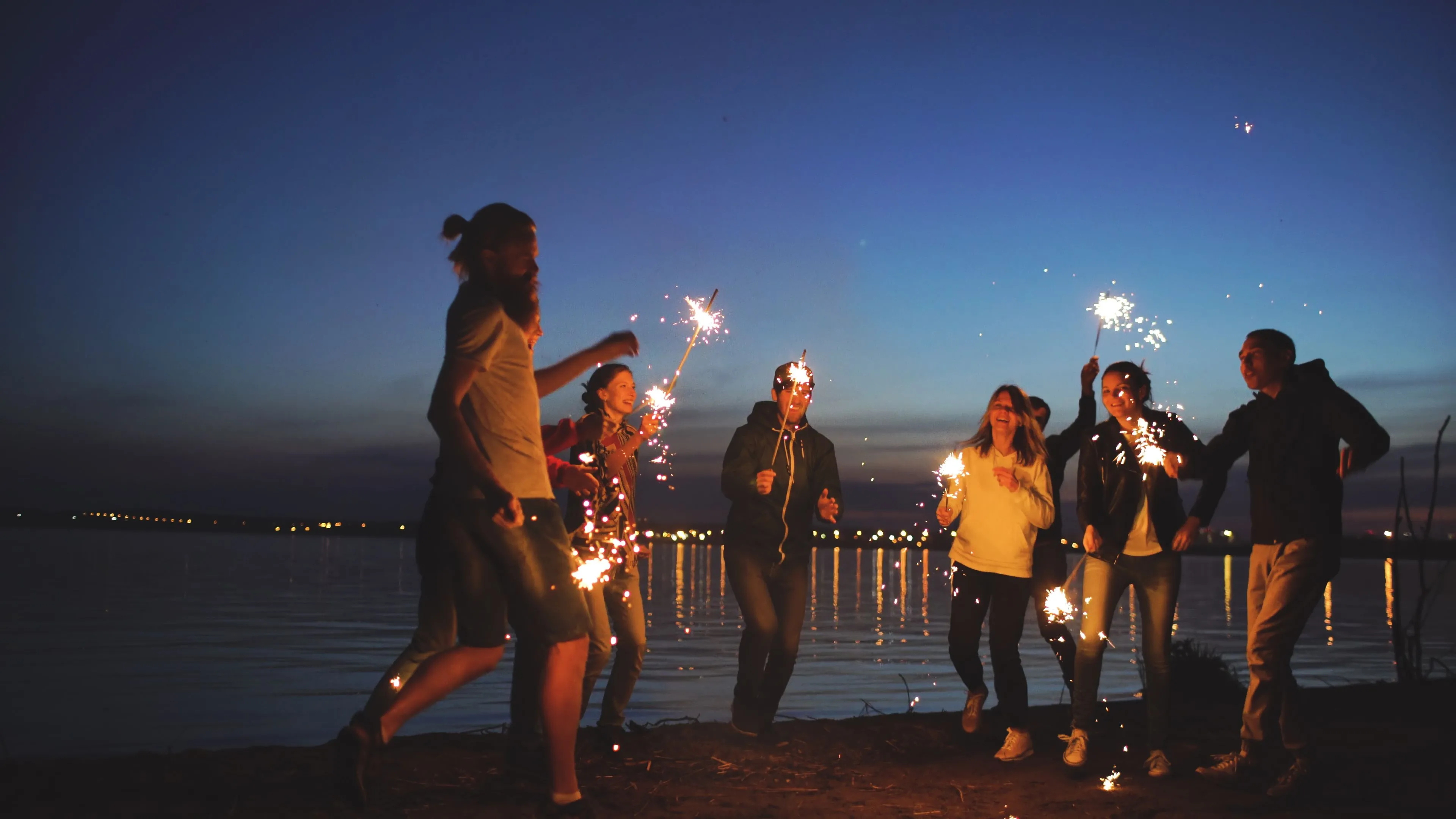 A group running along a lakeshore at dusk with sparklers, city lights strung across the water behind them
