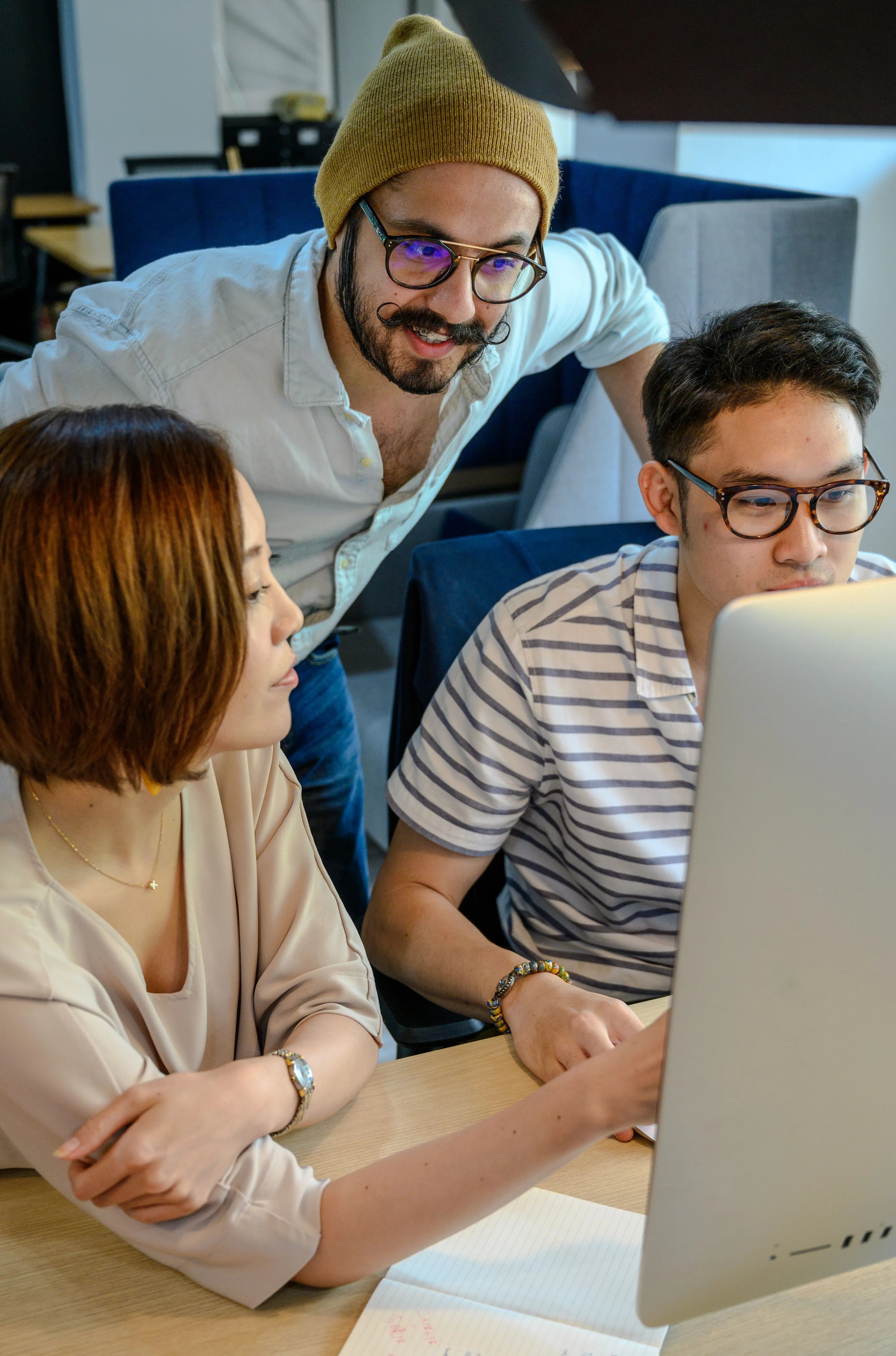 Three colleagues gathered around an iMac at a shared desk, talking through what is on the screen